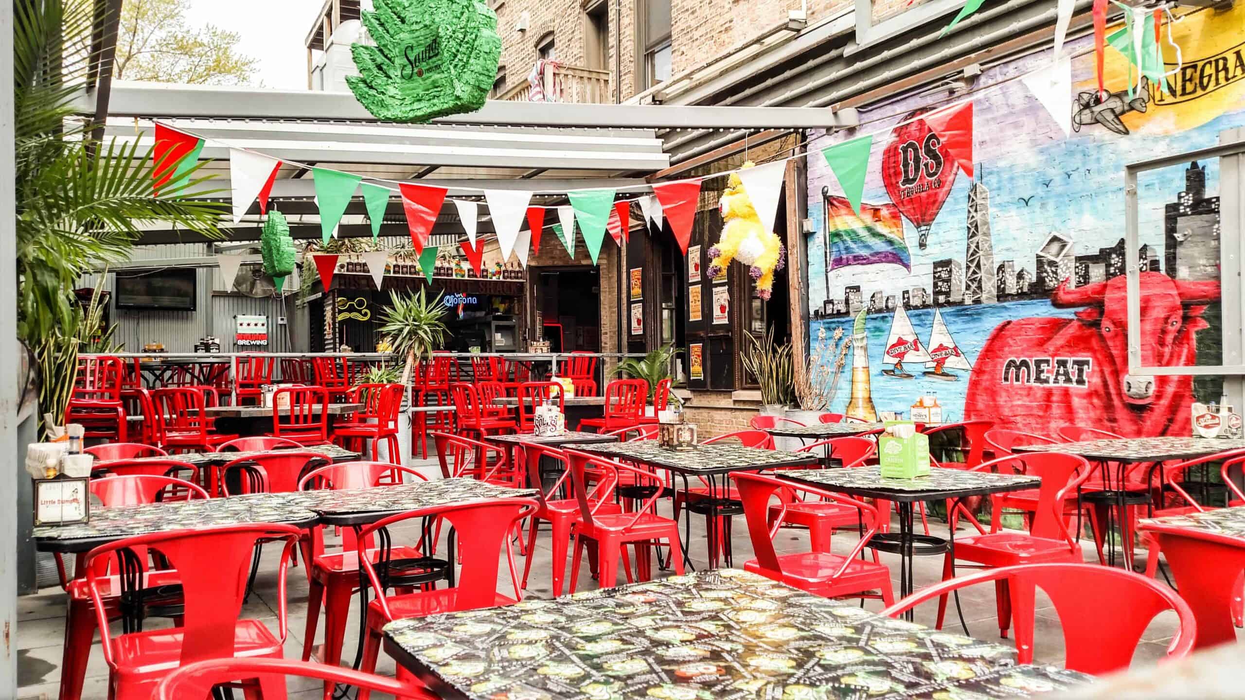 Bright outdoor dining space with colorful banners, mural art, and red chairs in Chicago’s North Halsted.